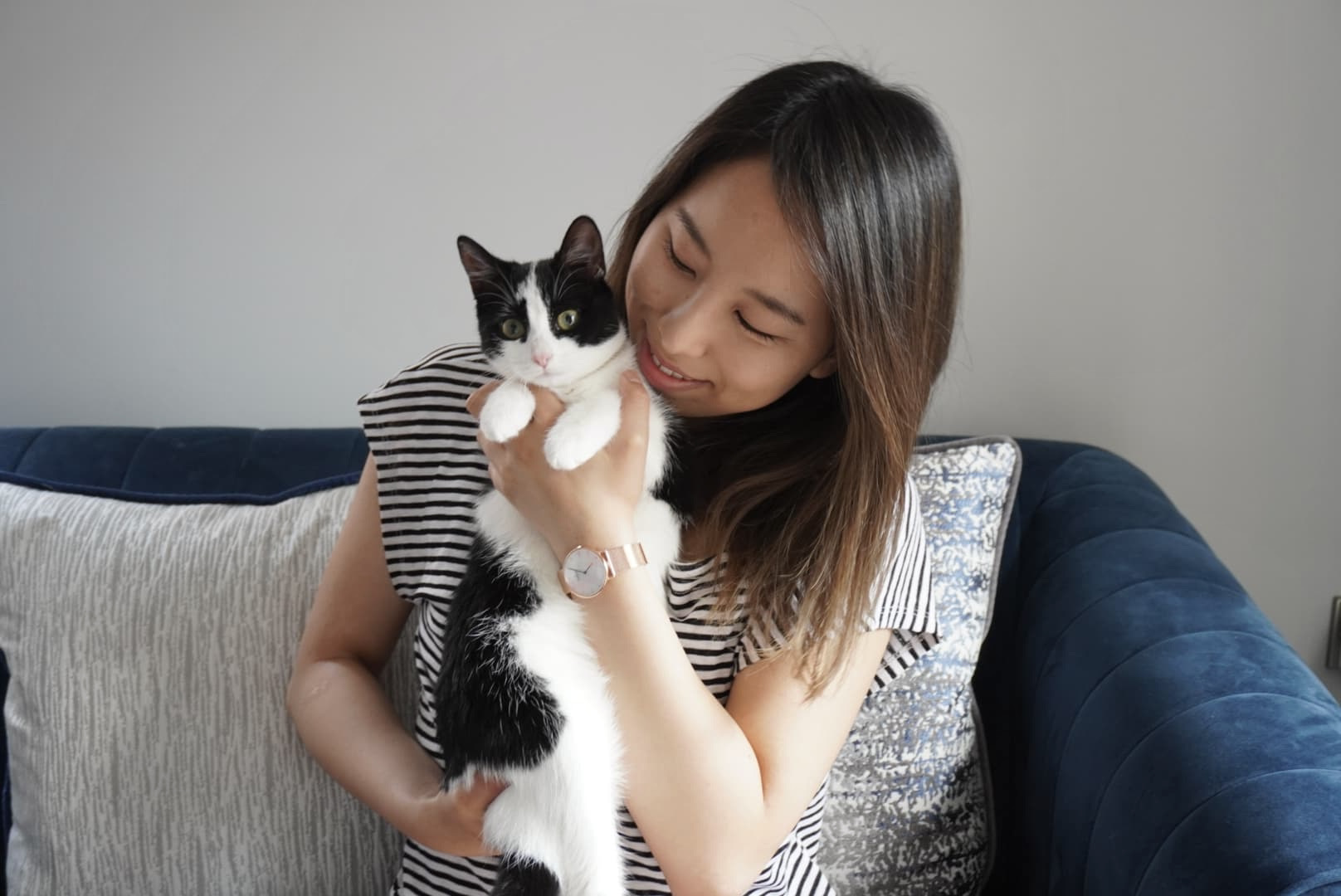 Portrait of Beverley Ho, vet content writer. A woman sits on a navy sofa. She is holding a black and white cat in her arms