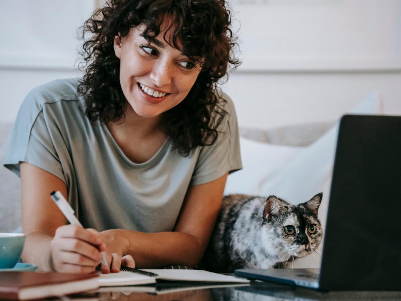 A cat writer with dark curly hair smiles at her laptop while writing notes in a book. She has a cat beside her